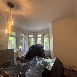 KrysTech - Pokój w trakcie przygotowań do odświeżenia: meble zabezpieczone czarną folią, drabina, nowoczesna lampa sufitowa z zakrytym kloszem. Jasne ściany i duże okna wykuszowe.