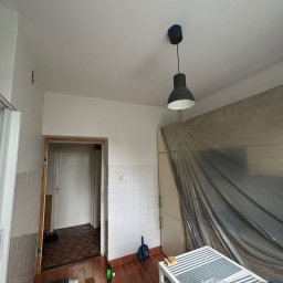 KrysTech - Pomieszczenie w trakcie prac remontowych: świeżo pomalowane białe ściany i sufit, podłoga z brązowo-szarych płytek, a meble zabezpieczone folią ochronną. Widoczne stare kafelki na ścianach.