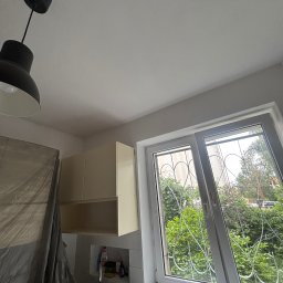 KrysTech - Świeżo pomalowany biały sufit z czarną lampą wiszącą, beżowe szafki kuchenne i uchylone okno z ozdobnymi kratami w kształcie serc. Część zlewu osłonięta folią.