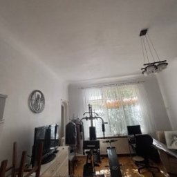 KrysTech - Jasny pokój z białymi ścianami i sufitem, drewnianą podłogą, dużym oknem z firankami oraz wiszącą lampą. W pomieszczeniu widoczny telewizor, sprzęt do ćwiczeń i biurko.