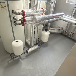 KrysTech - Nowoczesna instalacja grzewcza z dwoma białymi zbiornikami, rurami w srebrnej izolacji, czerwonymi zaworami, manometrami i naczyniami wzbiorczymi, na szarej posadzce.