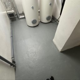 KrysTech - Pomieszczenie techniczne z dwoma białymi zbiornikami, szarą posadzką, widocznymi rurami instalacyjnymi i kanałem wentylacyjnym.