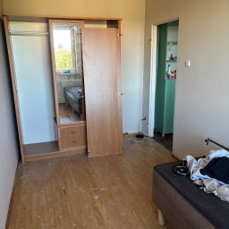 KrysTech - Pokój z drewnianą szafą z lustrem, podłoga z paneli pokryta resztkami farby i pyłu. Ściany w dwóch odcieniach beżu, w odbiciu lustra widać okno i grzejnik.