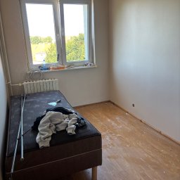 KrysTech - Jasny pokój z oknem, świeżo przygotowanymi ścianami i drewnianą podłogą z zabrudzeniami. Na łóżku leżą ubrania i drobne narzędzia, obok grzejnika.
