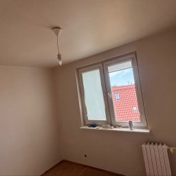 KrysTech - Świeżo wykończone pomieszczenie z beżowymi ścianami, oknem z widokiem na czerwony dach, prostą lampą i białym grzejnikiem panelowym pod parapetem.