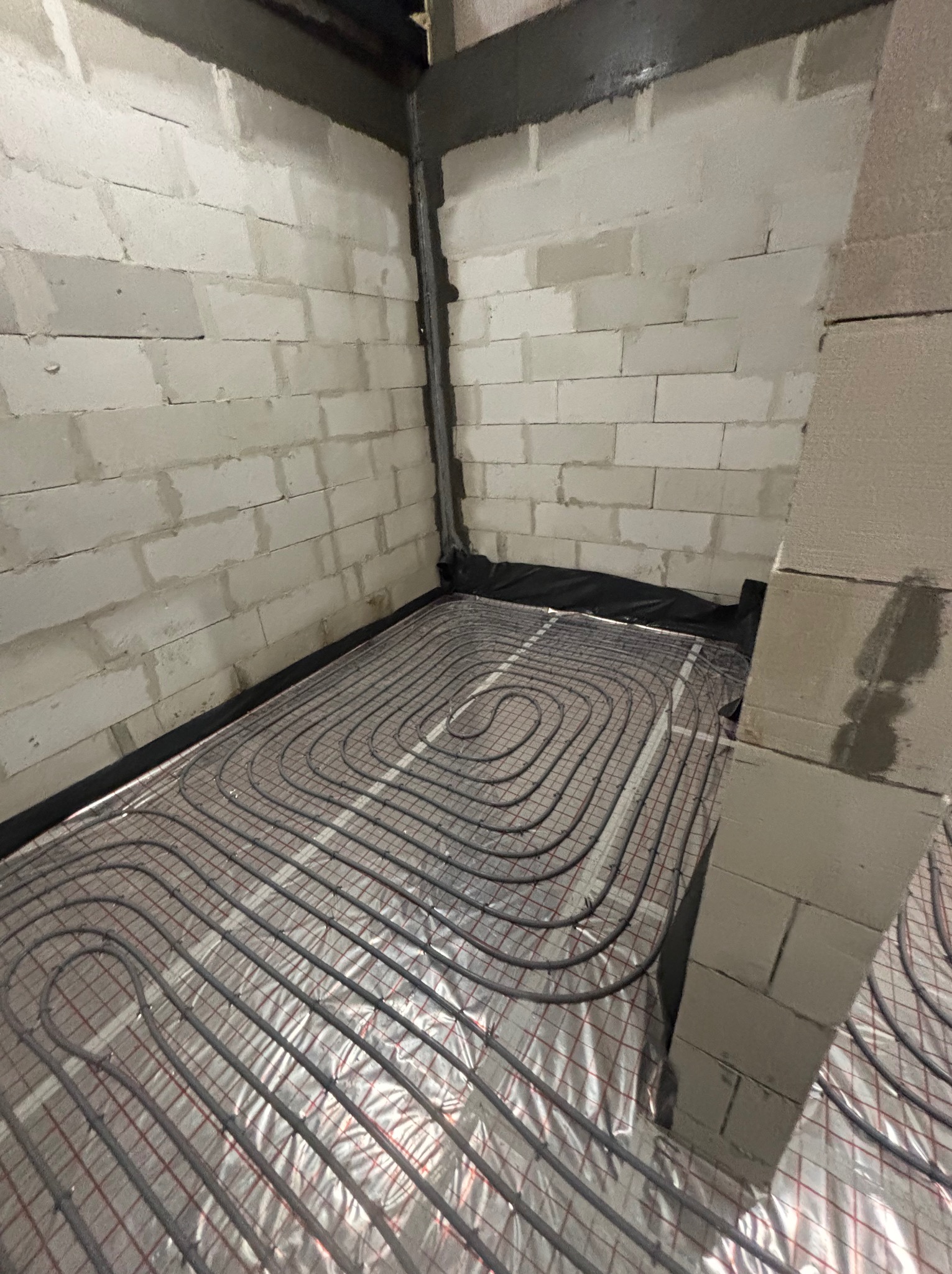 Ułożone rury ogrzewania podłogowego na folii izolacyjnej w surowym pomieszczeniu z betonowych bloczków. Widok z góry, instalacja przed wylaniem posadzki.