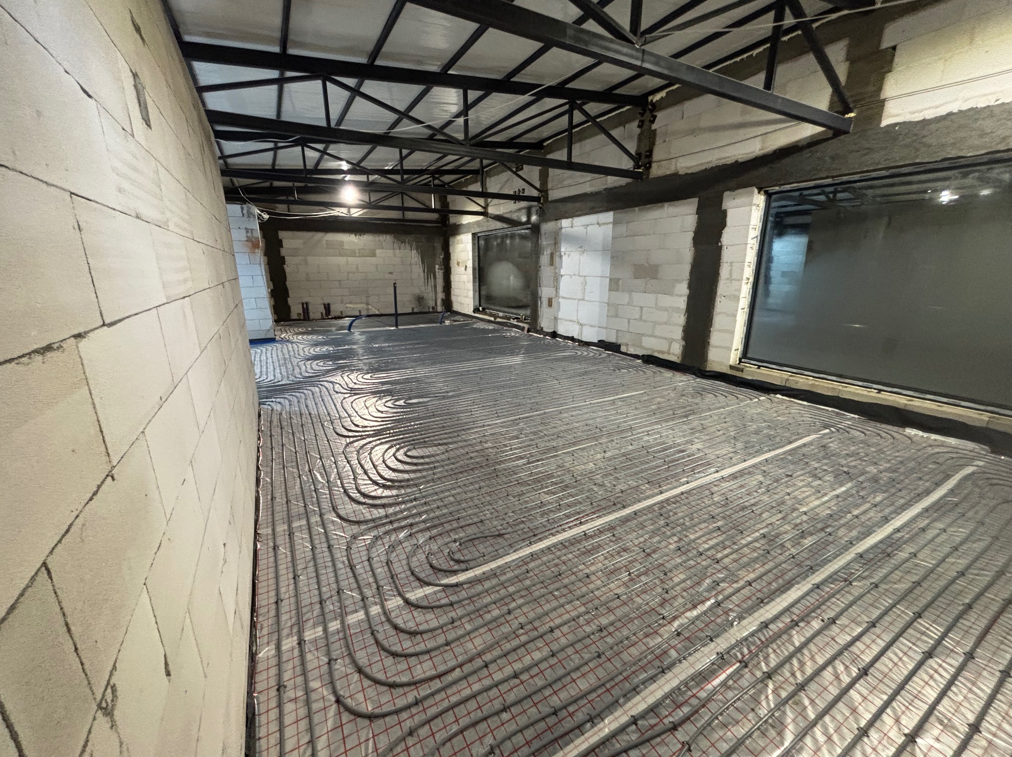 Szare rury ogrzewania podłogowego ułożone na folii z czerwoną siatką w surowym wnętrzu z białych bloczków i metalową konstrukcją sufitu. Widok z góry, szeroki kąt.