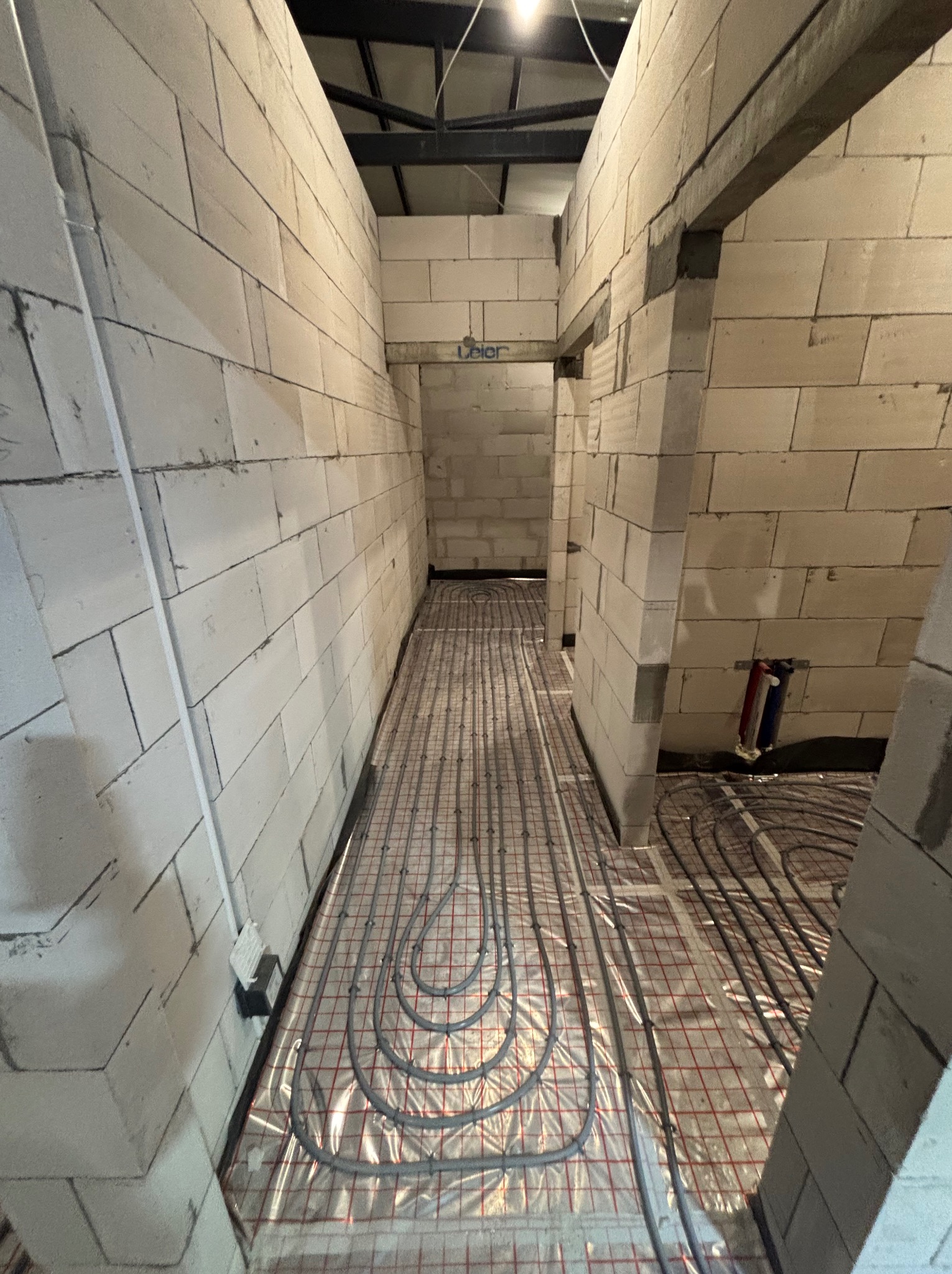 Montaż ogrzewania podłogowego w surowym wnętrzu. Szare rury ułożone na folii z czerwoną kratką, ściany z białych bloczków, widoczne instalacje hydrauliczne.