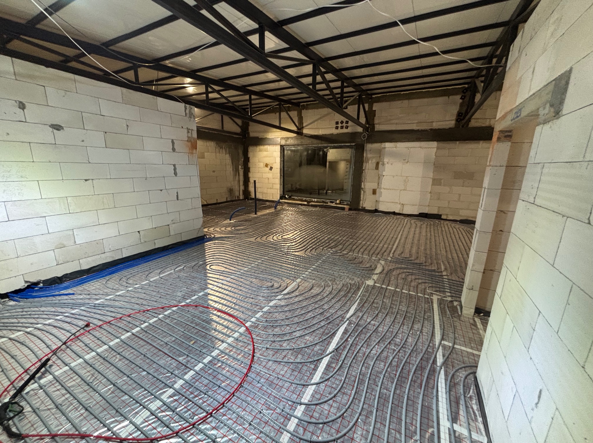 Szare rury ogrzewania podłogowego ułożone spiralnie na folii w surowym wnętrzu z białych bloczków i metalową konstrukcją sufitu. Widoczne niebieskie rury przy ścianie.