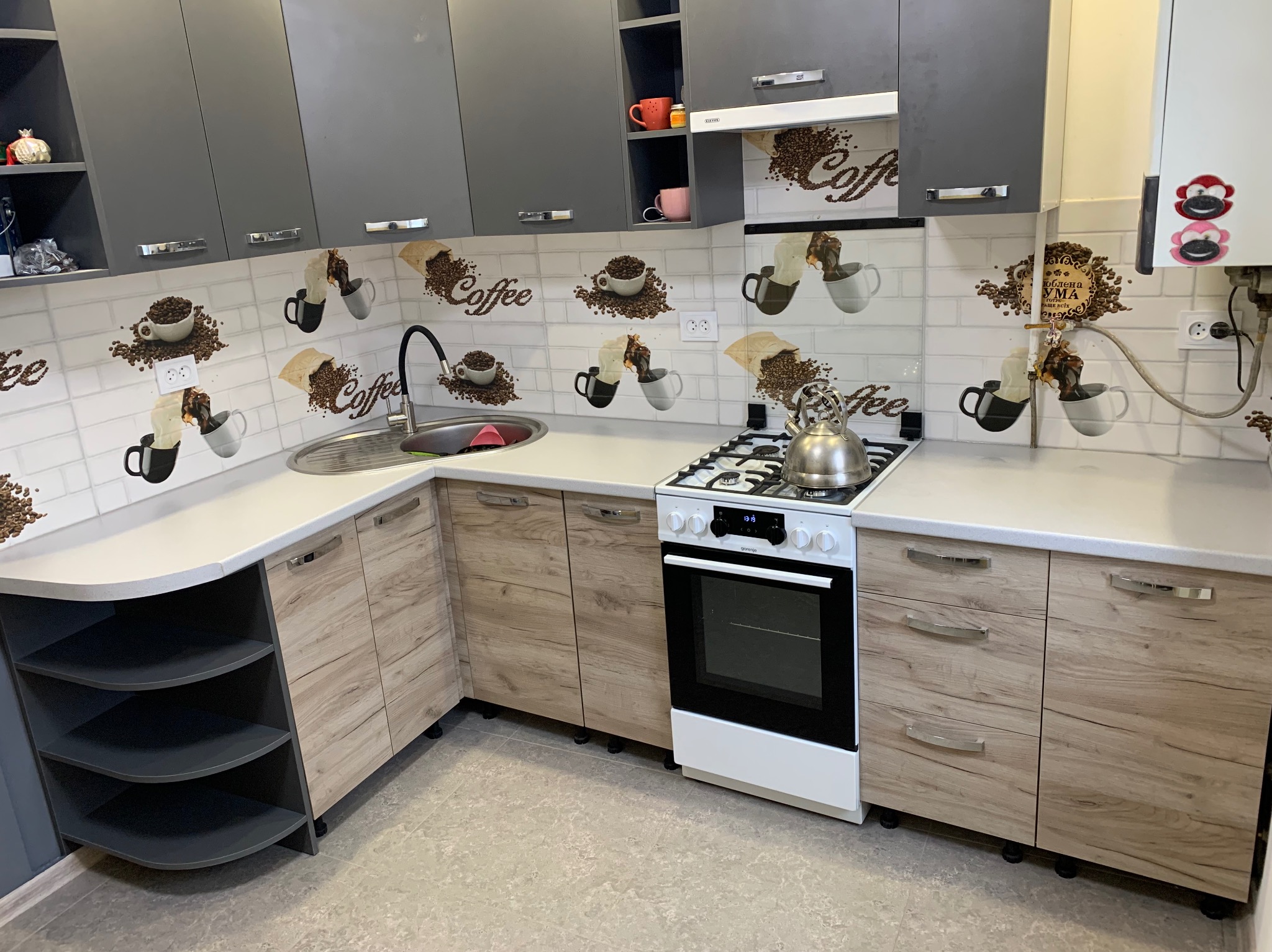 Nowoczesna kuchnia z szarymi szafkami górnymi i drewnopodobnymi dolnymi. Na ścianie płytki z motywem kawy. Biała kuchenka i czajnik na blacie.
