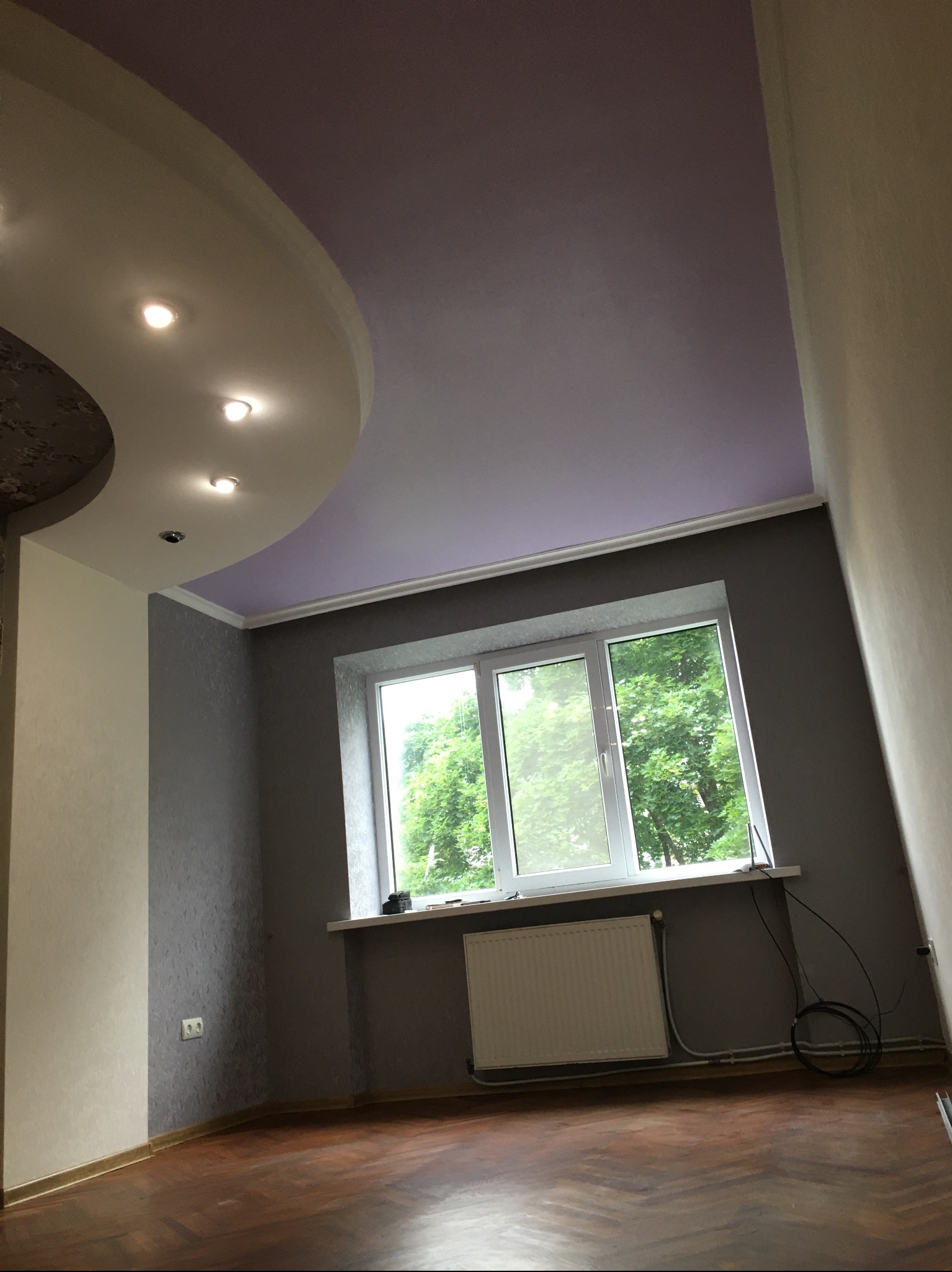 Pokój po remoncie: ściany w odcieniach szarości z fakturą, drewniana podłoga w jodełkę, okno z widokiem na zieleń, sufit z podwieszaną konstrukcją i oświetleniem punktowym.