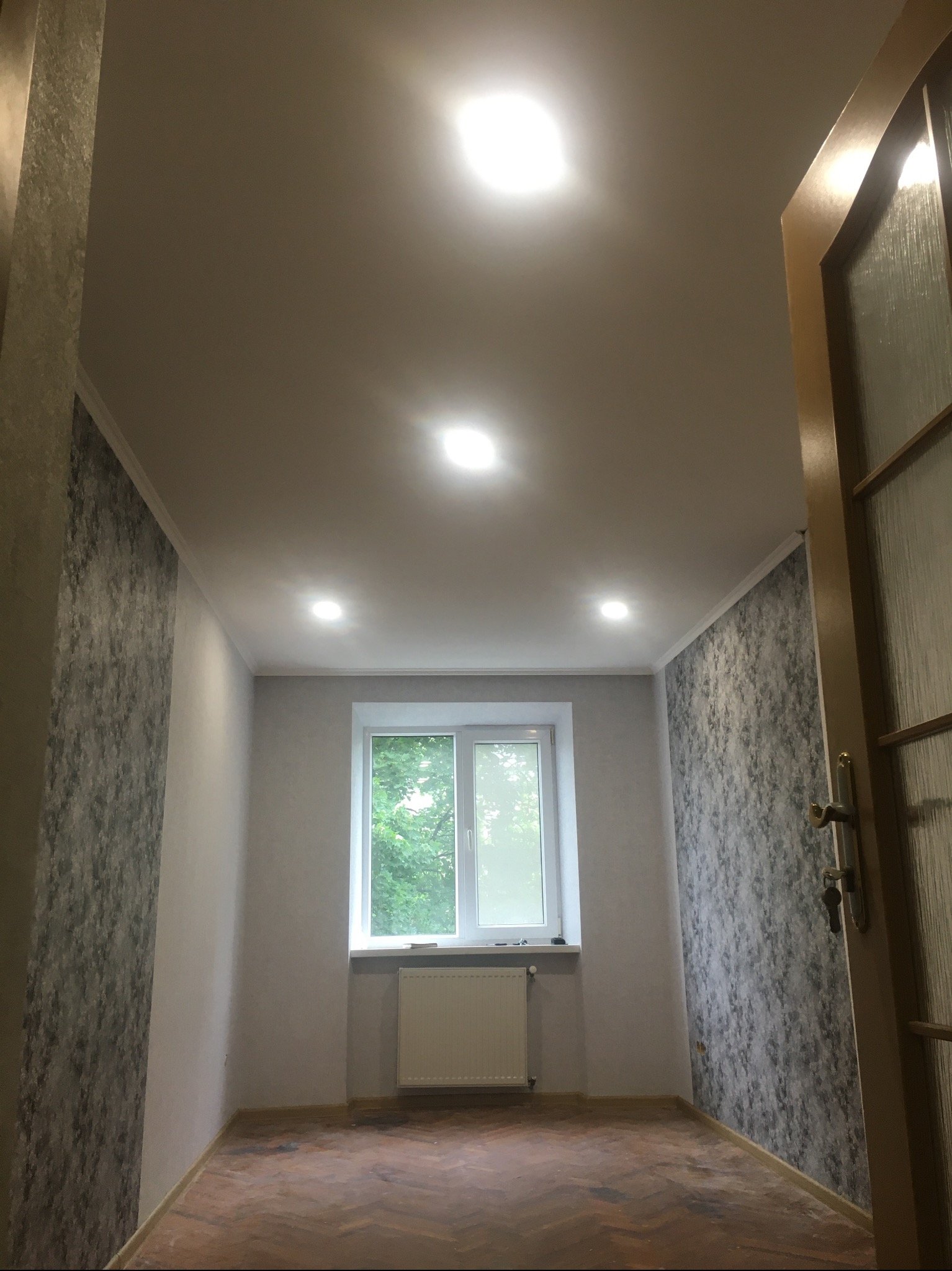 Pokój po remoncie: parkiet w jodełkę, ściany z tapetą w odcieniach szarości, białe okno z widokiem na zieleń, oświetlenie sufitowe i otwarte drzwi.