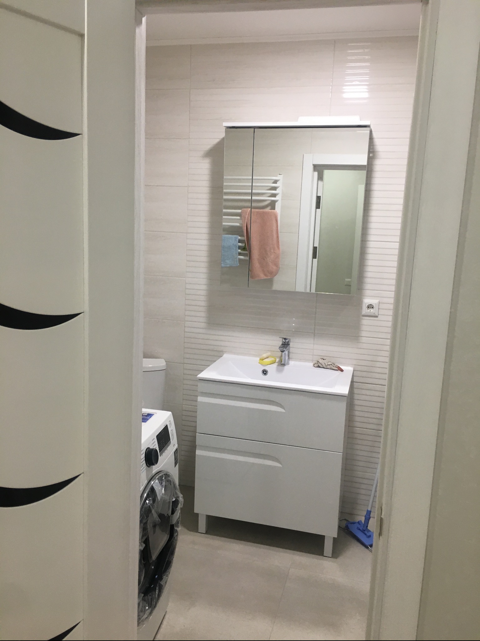 Jasna łazienka z umywalką w szafce, lustrem z szafką, pralką i mopem. Widok zza otwartych drzwi, ukazujący minimalistyczny design i funkcjonalne wyposażenie.