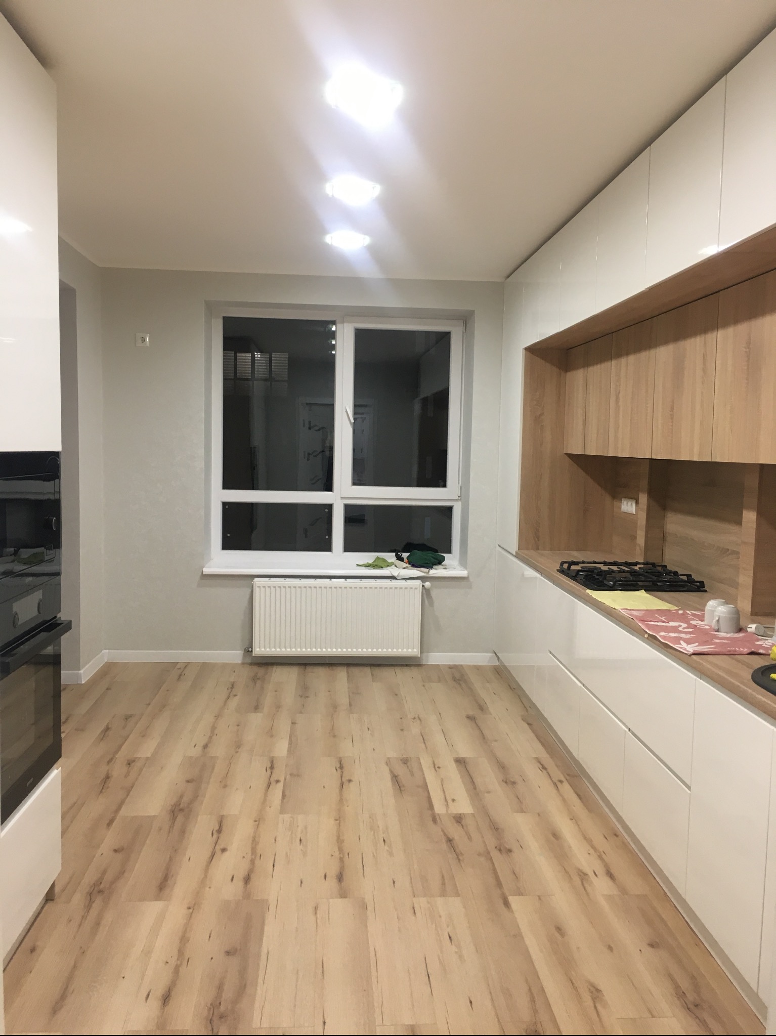 Nowoczesna kuchnia z białymi szafkami i drewnianym blatem. Widoczne okno z grzejnikiem i drewniana podłoga. Minimalistyczny design, jasne oświetlenie.