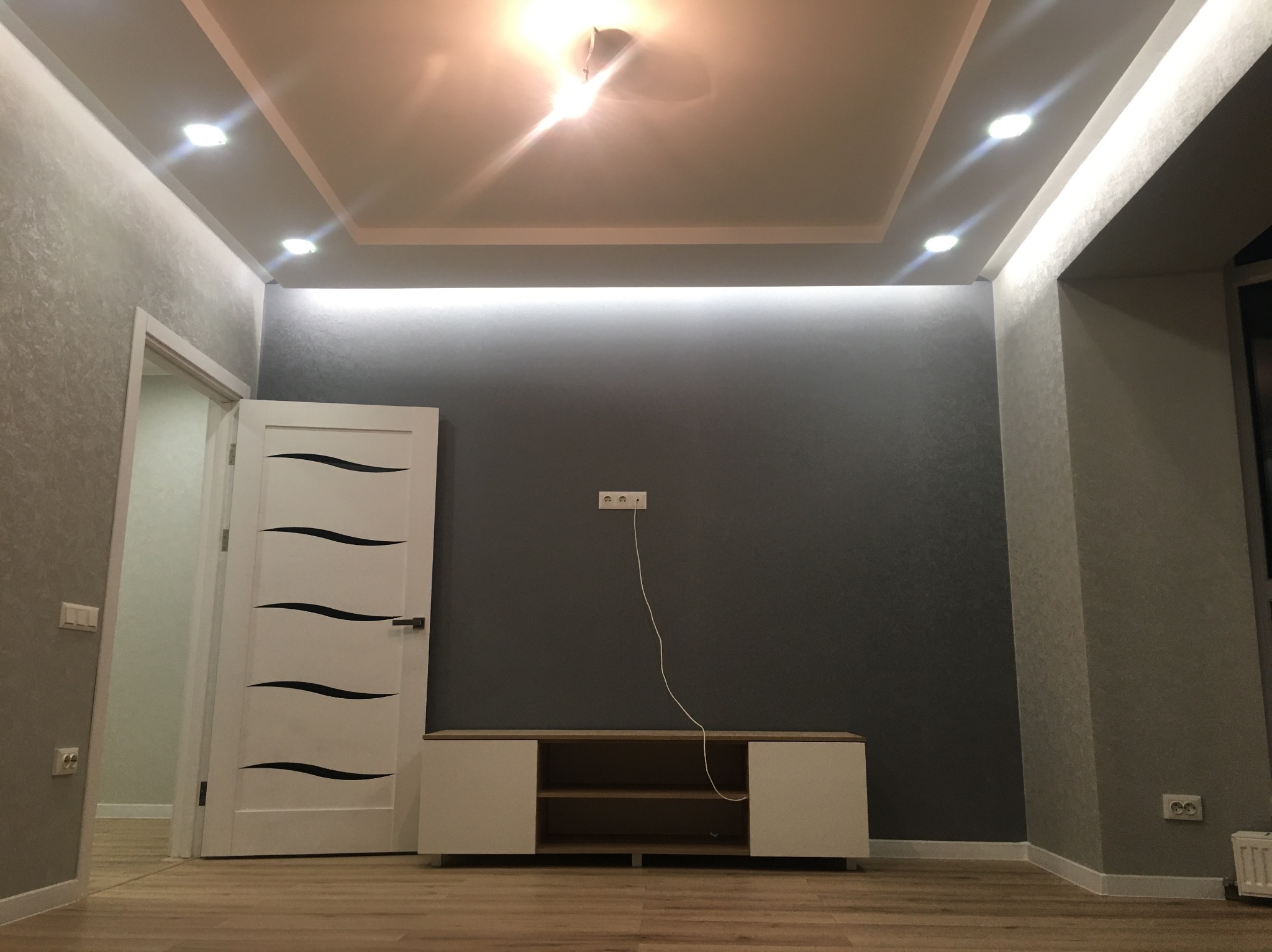Pokój po remoncie: szare ściany z fakturą, podwieszany sufit z oświetleniem LED, białe drzwi z dekoracyjnymi wstawkami i szafka RTV. Widok na odnowione wnętrze.
