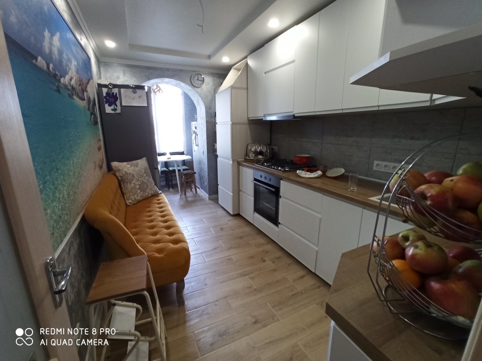 Nowoczesna kuchnia z białymi szafkami, drewnianym blatem i sofą w kolorze musztardowym. Na ścianie fototapeta z motywem morskim. Podłoga wyłożona płytkami imitującymi drewno.