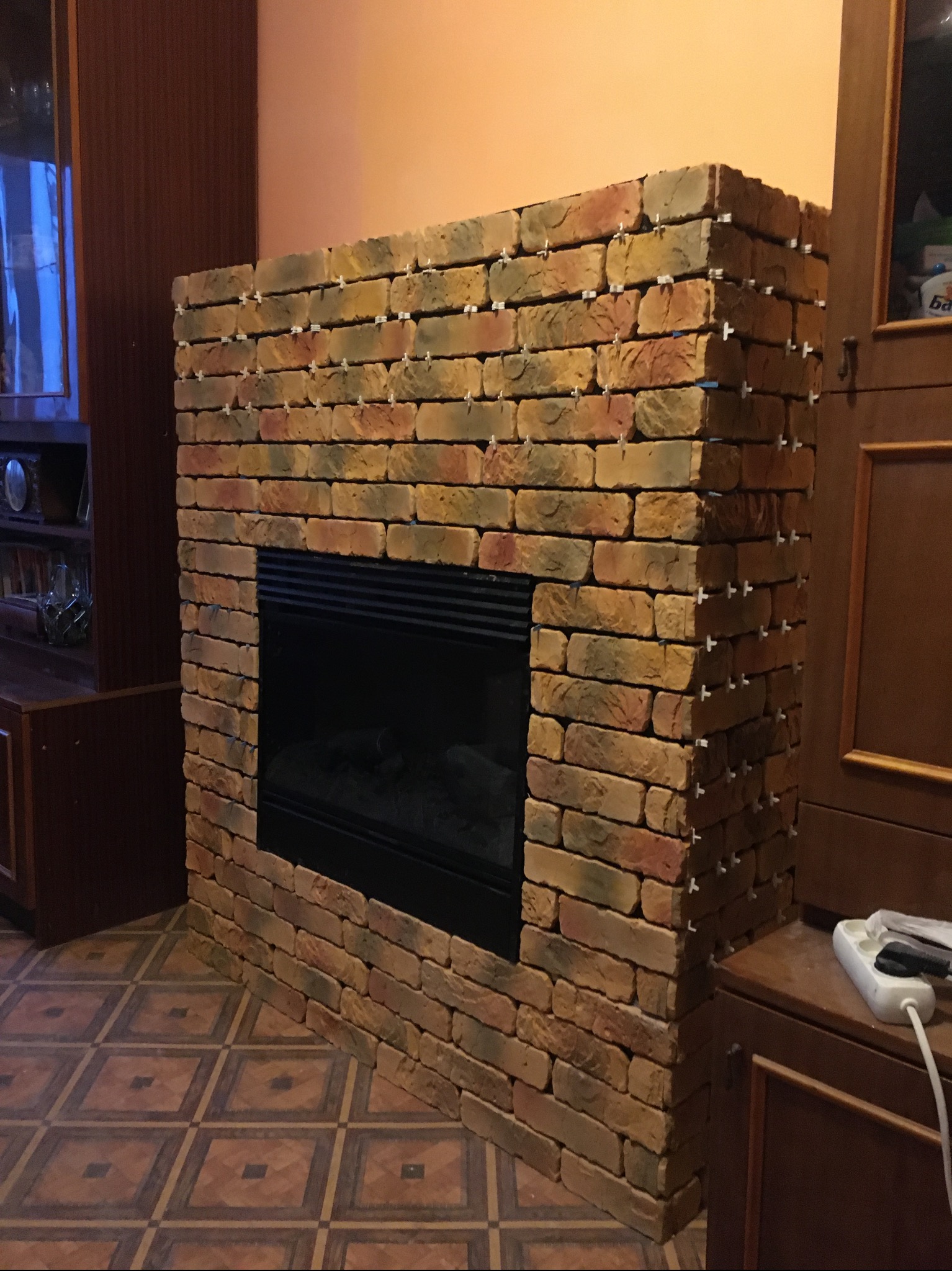 Wnętrze z kominkiem w trakcie obkładania cegłą klinkierową. Widoczne krzyżyki dystansowe. Cegła w odcieniach brązu i beżu. Na podłodze wzorzyste płytki.