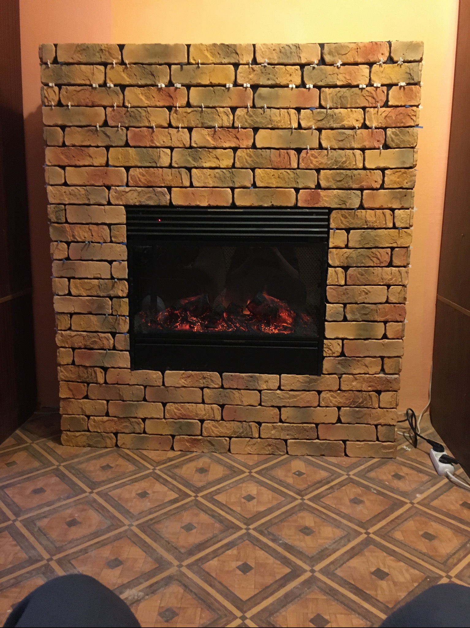 Ozdobny kominek z cegły klinkierowej z imitacją ognia, widoczny na tle wzorzystej podłogi i ściany w ciepłych barwach. Fragment nóg osoby w dolnej części kadru.