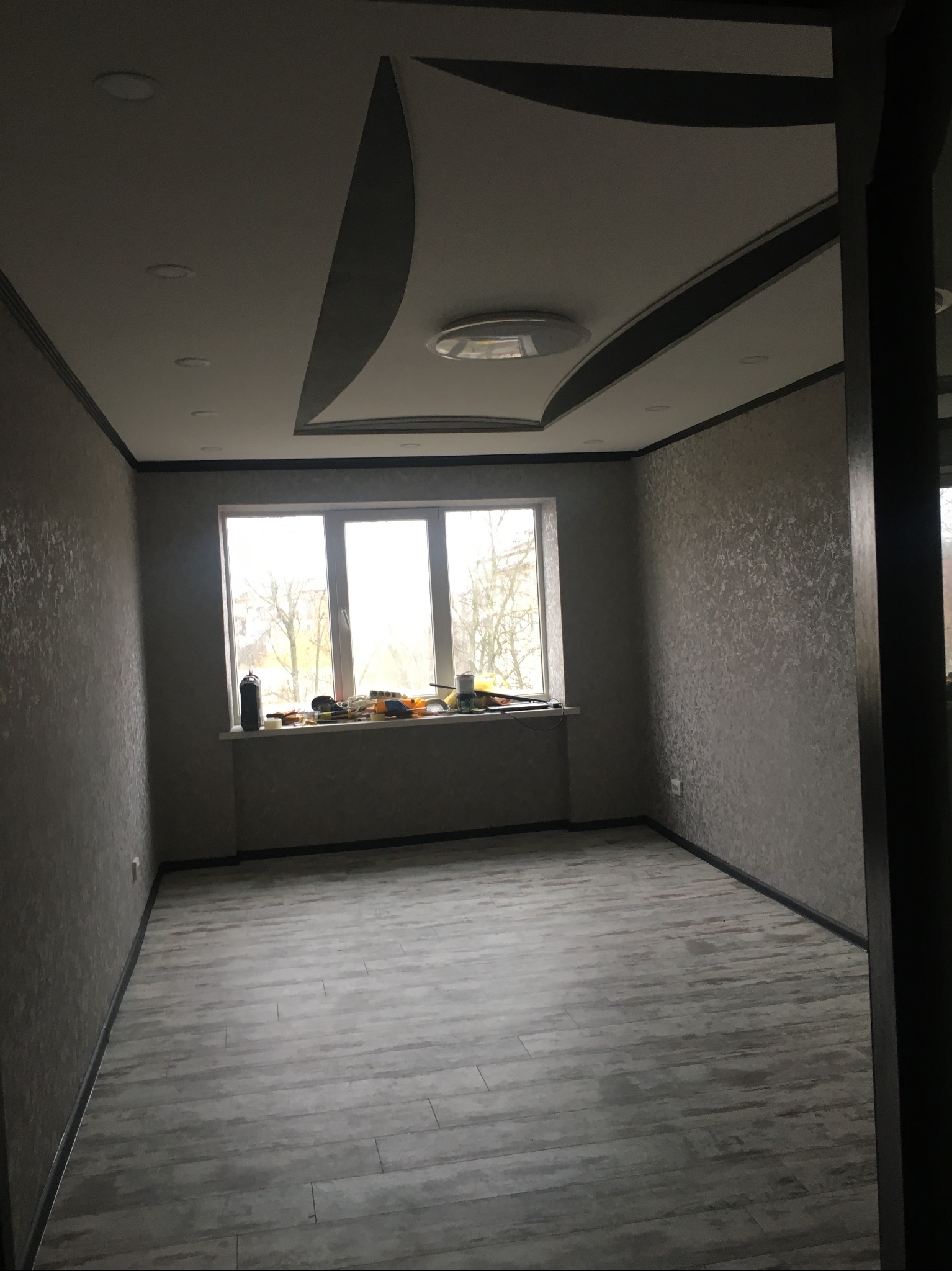 Wykończone wnętrze pokoju po remoncie: szare ściany z fakturą, panele podłogowe, okno z narzędziami na parapecie, ozdobny sufit podwieszany z oświetleniem.