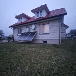 MW Docieplenia Podlasie - Dom z białego bloczka z czerwonym dachem i panelami słonecznymi na elewacji. Widoczne okna, antena satelitarna i trawnik. Pochmurne niebo w tle.