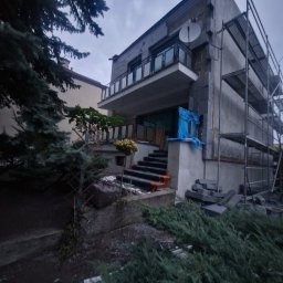 MW Docieplenia Podlasie - Dwupiętrowy dom w trakcie prac elewacyjnych, z widocznym rusztowaniem i materiałami budowlanymi. Część elewacji zabezpieczona folią. Ujęcie z dołu, pochmurne niebo.