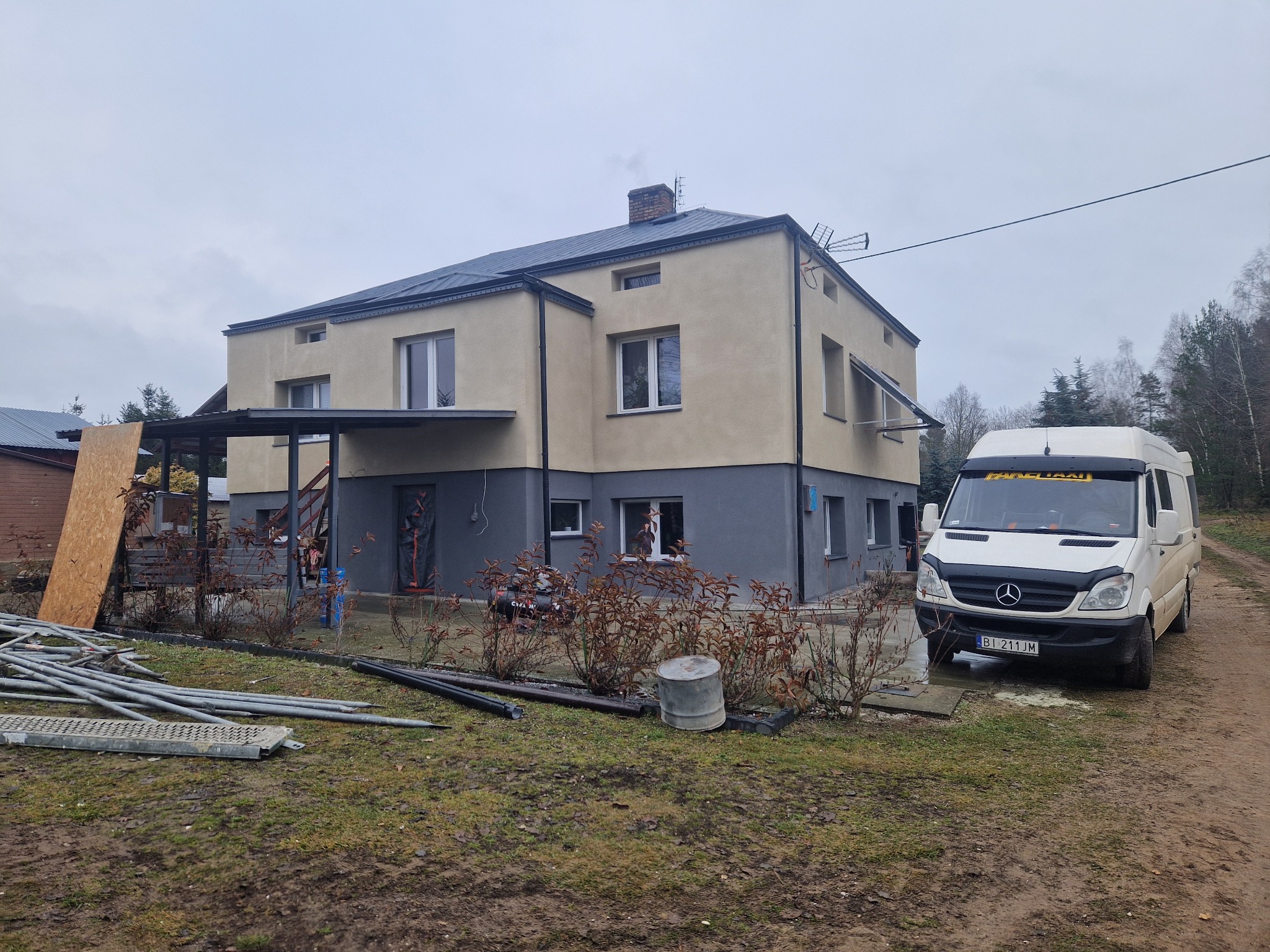 Dwukondygnacyjny dom z elewacją w odcieniach beżu i szarości, z widocznym samochodem dostawczym i elementami rusztowania na trawniku. Pochmurne niebo w tle.