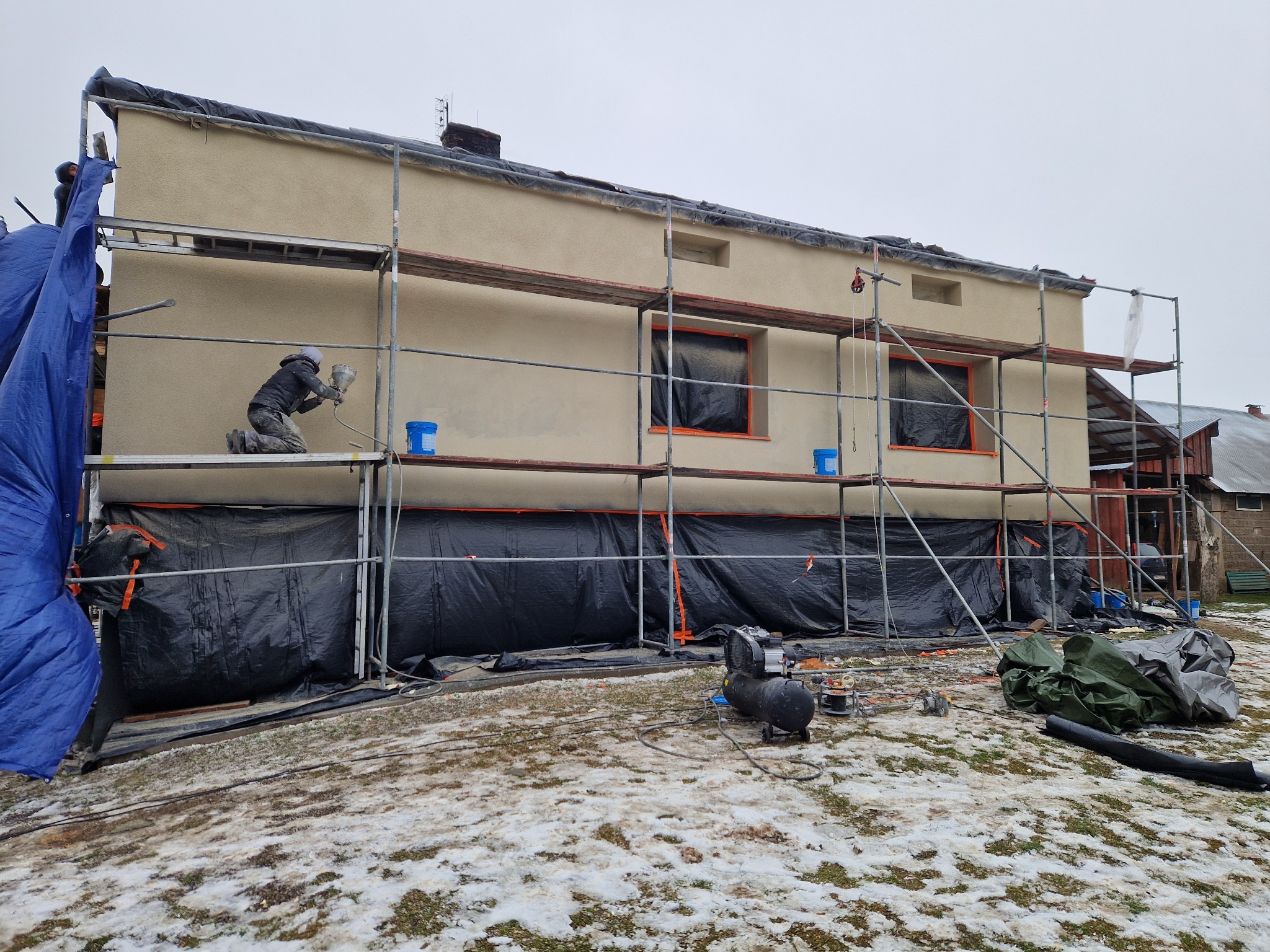 Pracownik w kombinezonie maluje elewację natryskowo z rusztowania. Okna zabezpieczone folią, widoczny kompresor i narzędzia. Częściowo ośnieżony teren.
