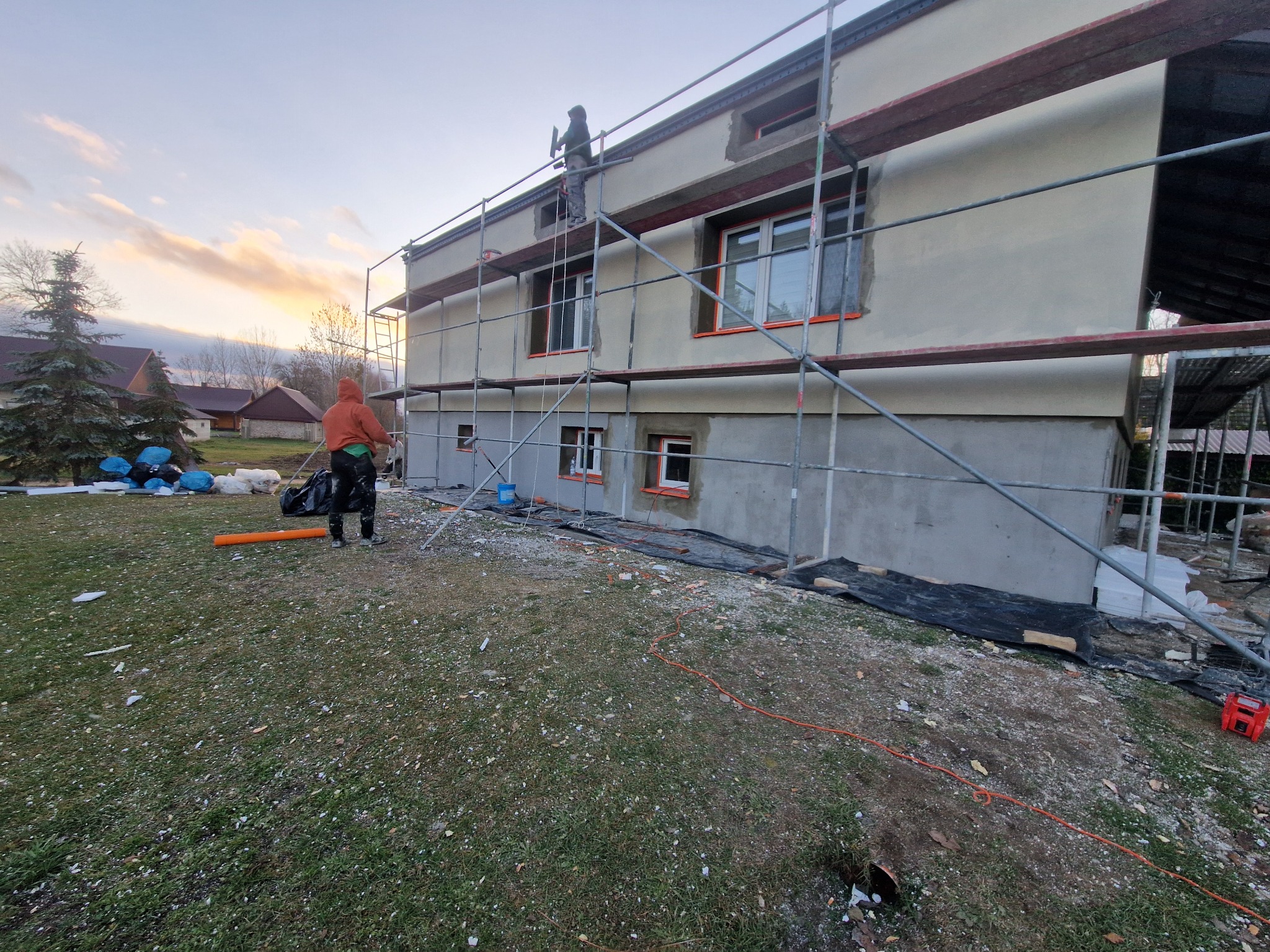 Dwukondygnacyjny budynek w trakcie prac elewacyjnych, otoczony rusztowaniami. Pracownik na rusztowaniu, widoczne materiały budowlane i narzędzia na trawniku.