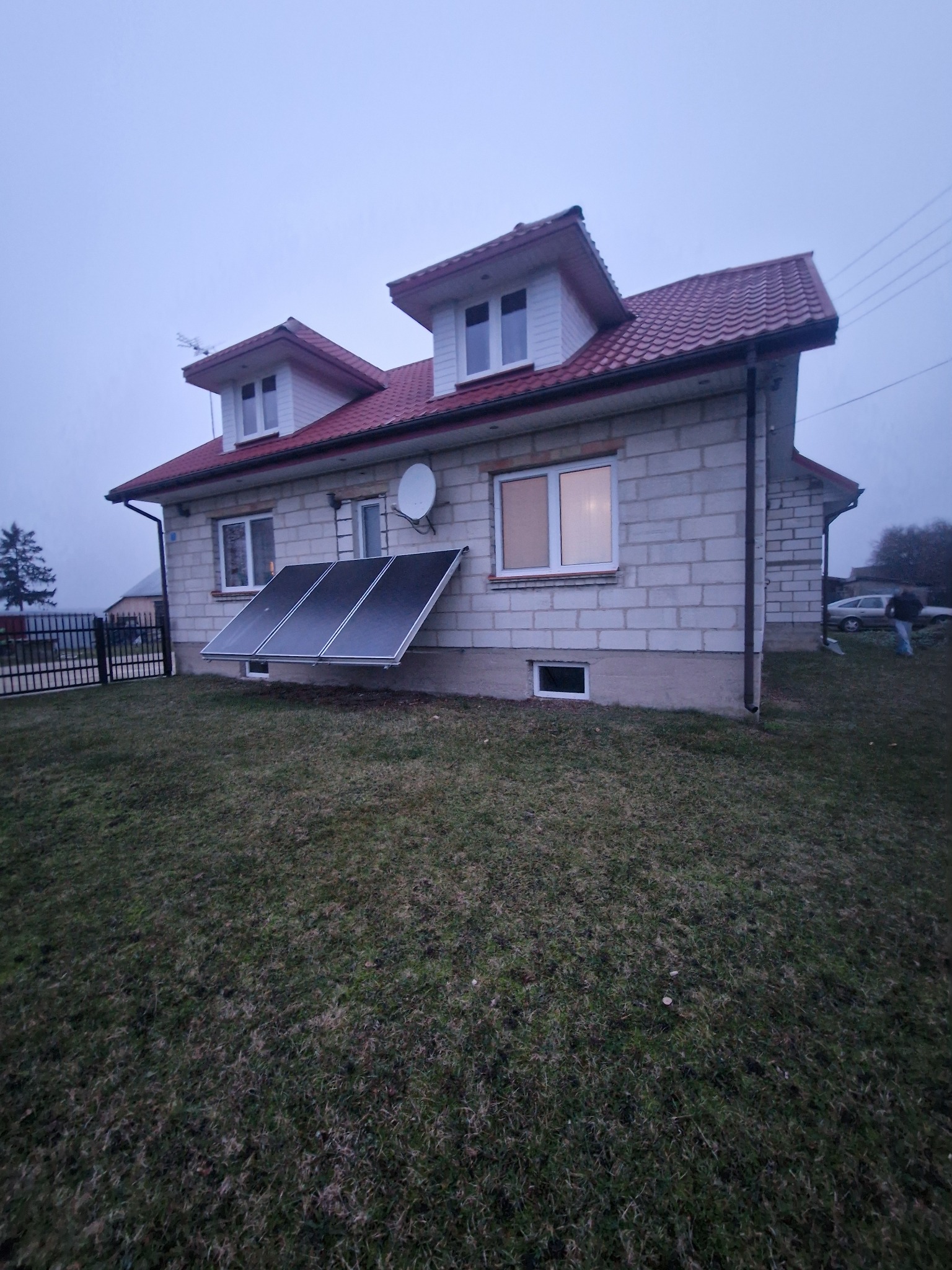 Dom z białego bloczka z czerwonym dachem i panelami słonecznymi na elewacji. Widoczne okna, antena satelitarna i trawnik. Pochmurne niebo w tle.