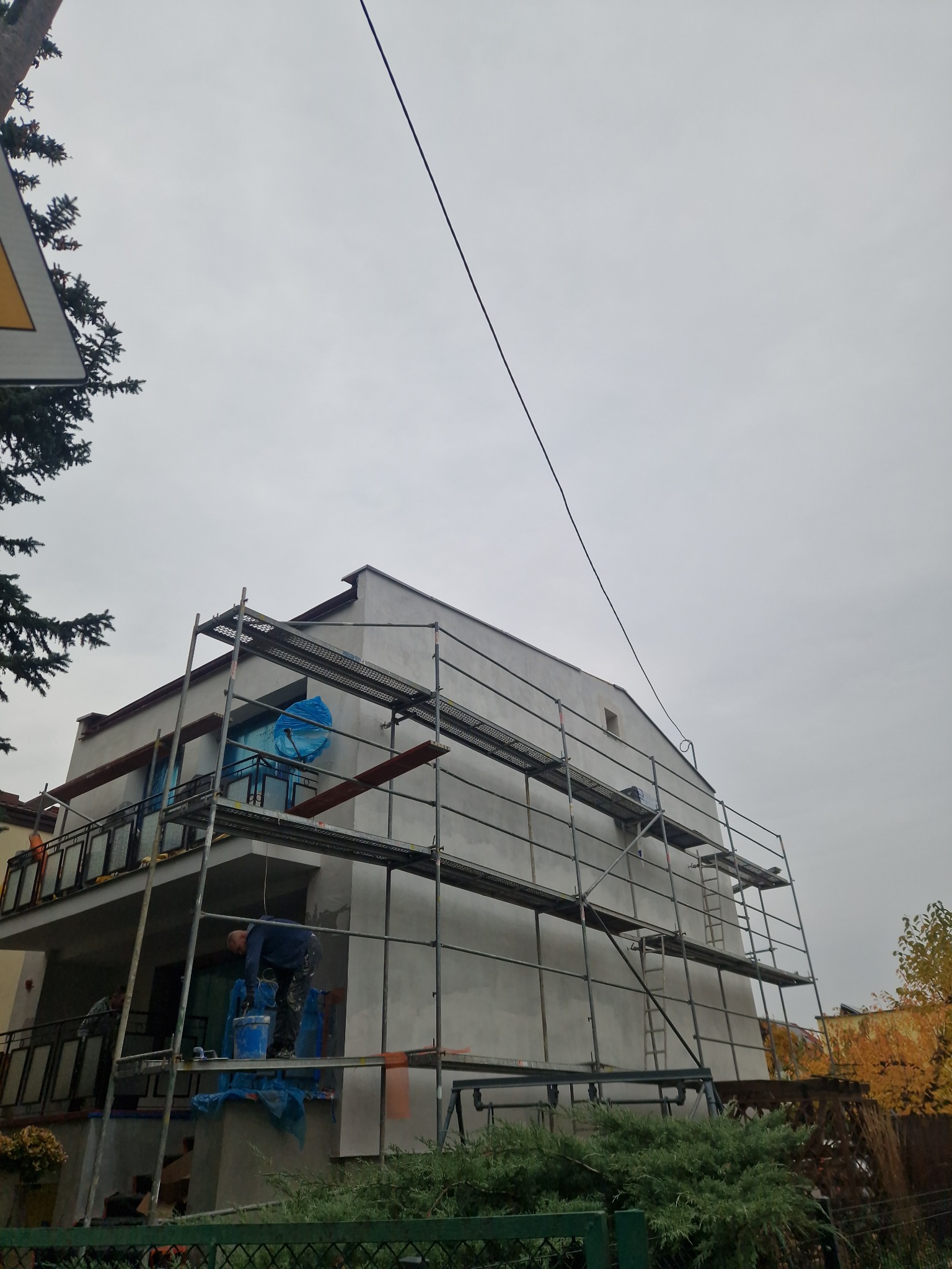 Dwupiętrowy dom w trakcie prac elewacyjnych, otoczony rusztowaniem. Widoczny pracownik przy pracy, zabezpieczone okna folią. Pochmurne niebo w tle.