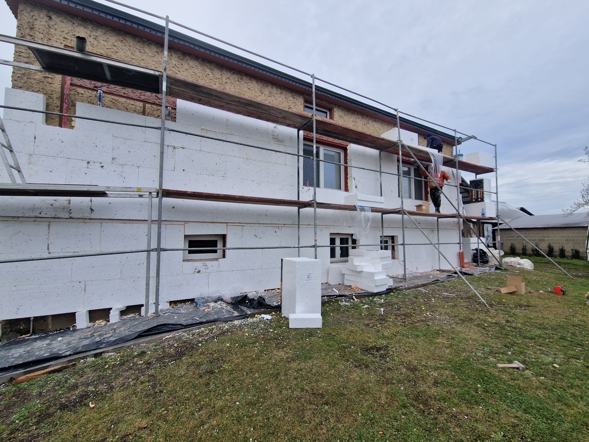 Dom w trakcie ocieplania styropianem, widoczne rusztowania i pracownicy. Częściowo ocieplona elewacja, odsłonięty fragment starej elewacji. Prace w toku.