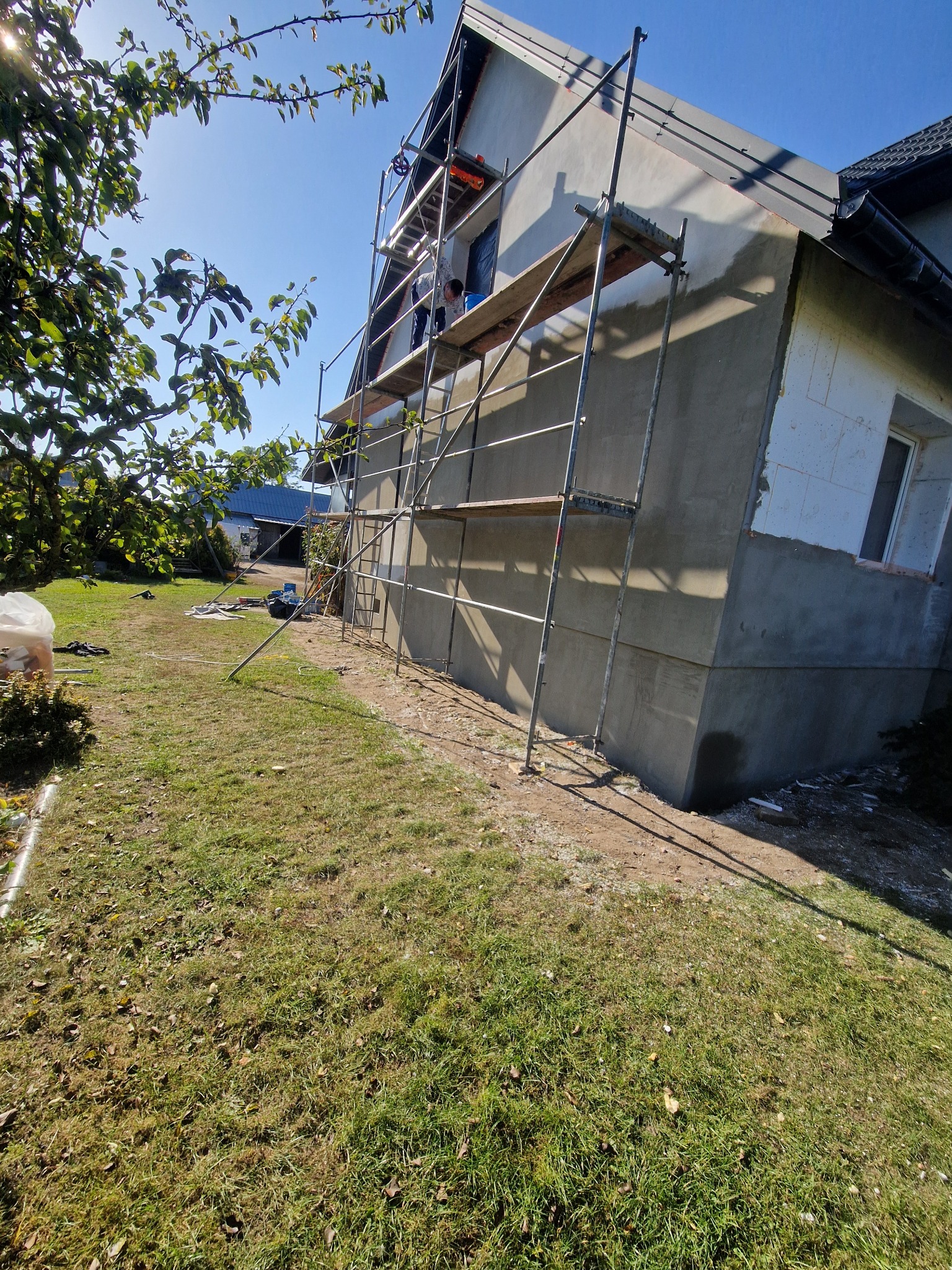 Dom w trakcie tynkowania elewacji, widoczne rusztowania i pracownik. Część ściany ocieplona styropianem, reszta pokryta szarym tynkiem. Słoneczny dzień.