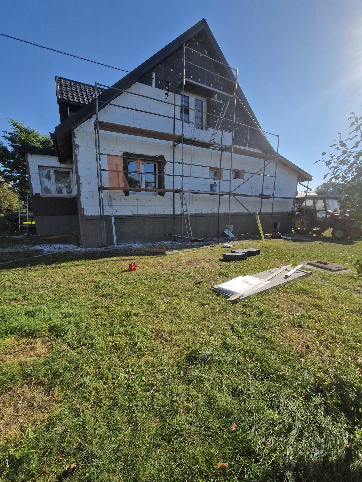 Dom w trakcie ocieplania: styropian na ścianach, rusztowania, częściowo otynkowany cokół. Na trawniku narzędzia i materiały budowlane, w tle traktor. Słoneczny dzień.