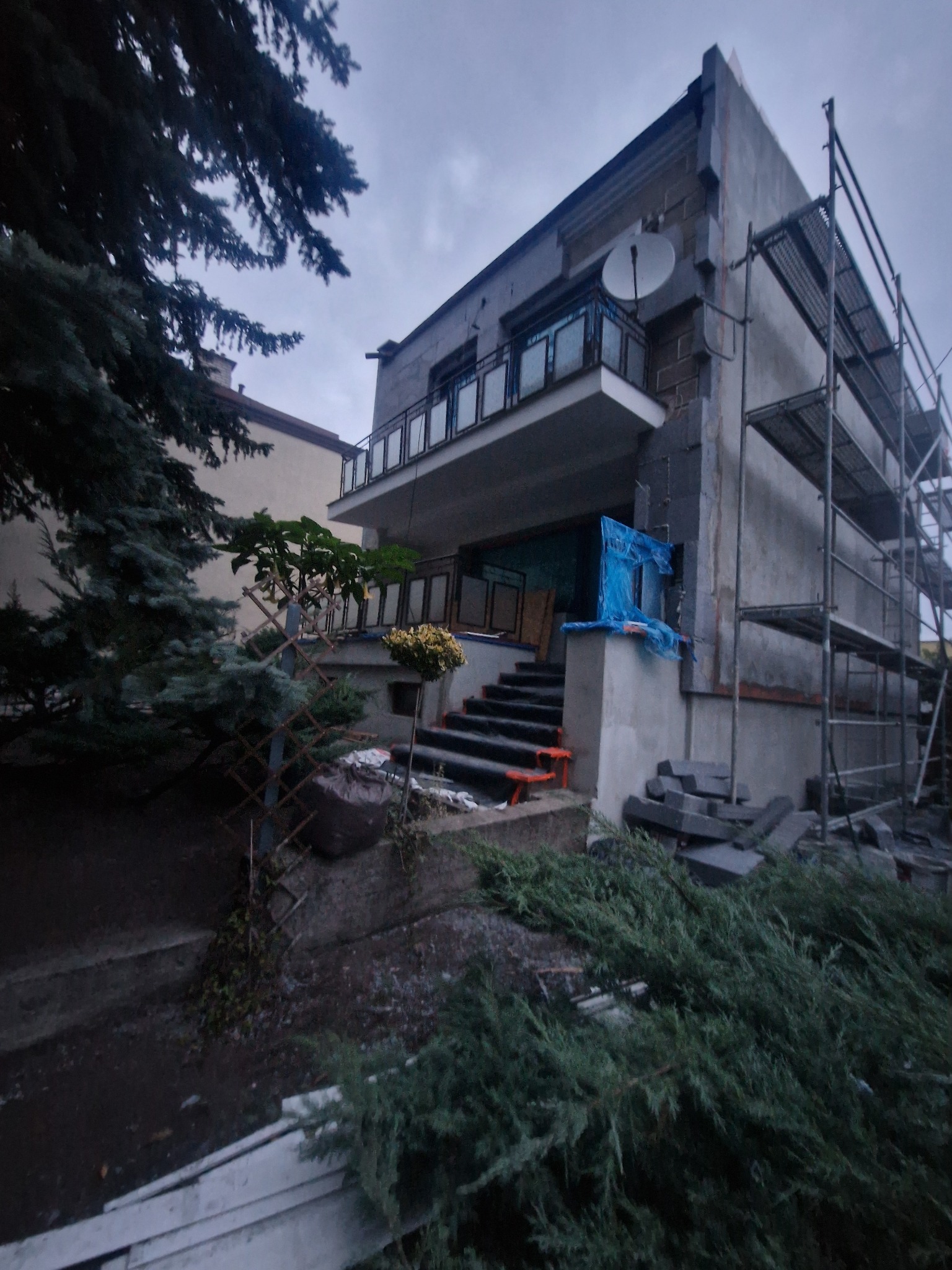 Dwupiętrowy dom w trakcie prac elewacyjnych, z widocznym rusztowaniem i materiałami budowlanymi. Część elewacji zabezpieczona folią. Ujęcie z dołu, pochmurne niebo.