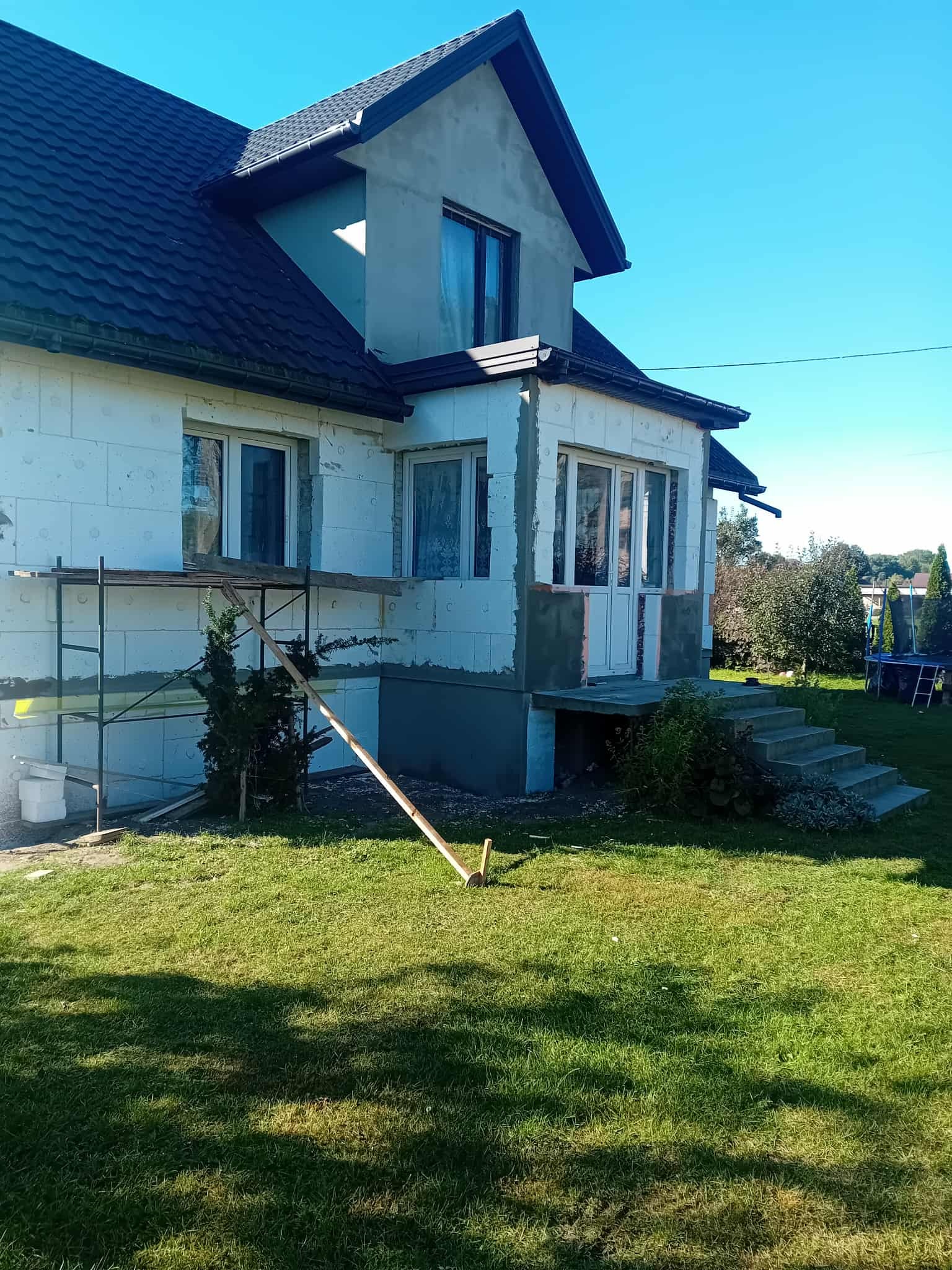 Dom jednorodzinny w trakcie prac elewacyjnych: ocieplenie styropianem, częściowo otynkowany, z rusztowaniem i elementami przygotowawczymi na trawniku.