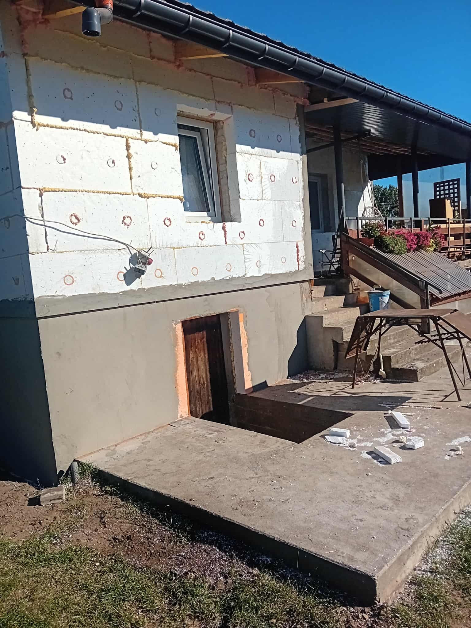 Dom w trakcie ocieplania styropianem, widoczne otwory po kołkach i szpary wypełnione pianką. Część ściany pomalowana na szaro. Wejście do piwnicy.