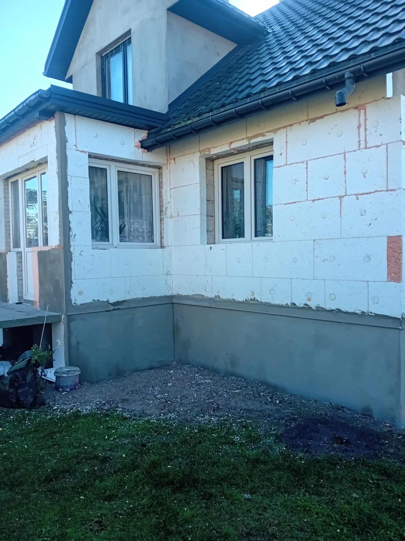 Niedokończona elewacja domu z białych bloczków, widoczne okna z zasłonami, szara podmurówka i fragment trawnika. Etap prac przed tynkowaniem.