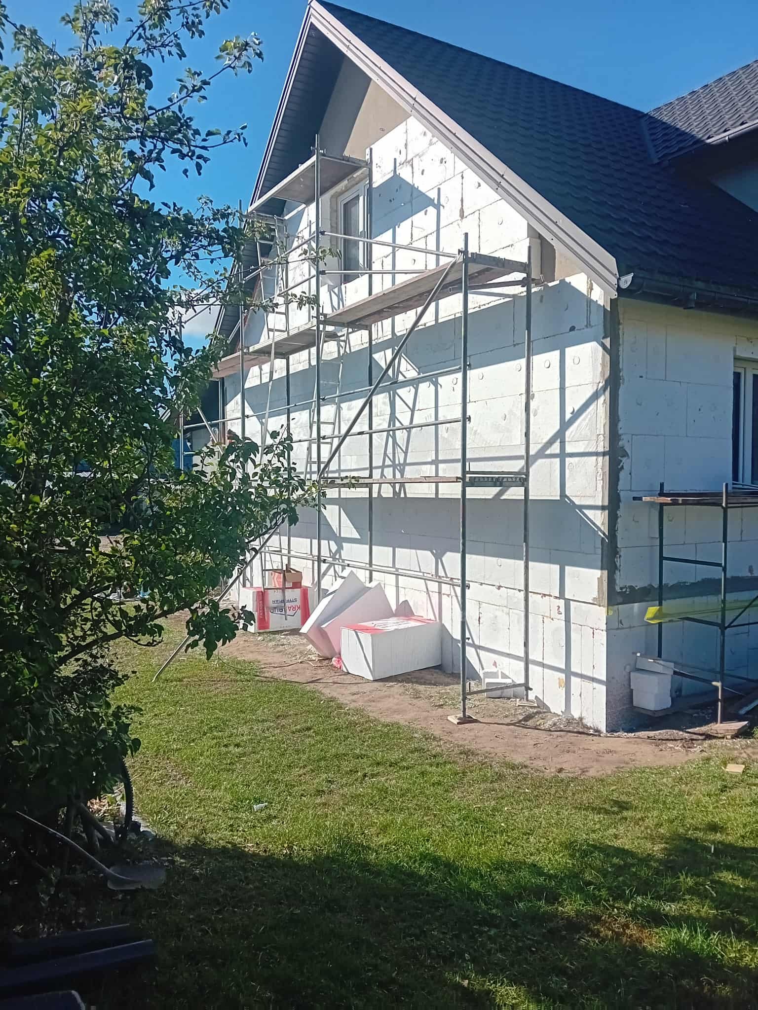 Dom w trakcie ocieplania, widoczne rusztowania, białe bloczki i styropian. Częściowo otynkowana ściana szczytowa. Słoneczny dzień, zielony trawnik.