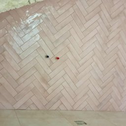 Usługi remontowe - Ściana wyłożona różową, błyszczącą glazurą w jodełkę, z widocznymi przyłączami wodnymi. Fragment sufitu z izolacją i podłoga z płytek w odcieniach beżu.