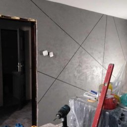 Usługi remontowe - Ściana z dekoracyjnym tynkiem w odcieniach szarości, zdobiona czarnymi liniami tworzącymi geometryczny wzór. Widoczne narzędzia i materiały budowlane.