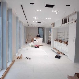 Usługi remontowe - Wnętrze sklepu w trakcie wykańczania: białe ściany, podłoga z płytek, zamontowane regały i oświetlenie. Widoczne narzędzia i materiały budowlane.