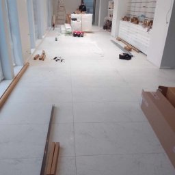 Usługi remontowe - Wnętrze sklepu w trakcie wykańczania: białe płytki podłogowe, regały z szufladami, narzędzia i materiały budowlane. Widoczna drabina i osoba w tle.