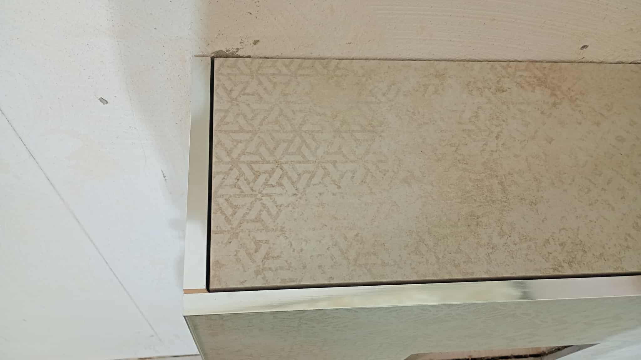 Beżowa płytka z geometrycznym wzorem, wykończona metalową listwą, zamontowana na ścianie. Widoczne niedoskonałości ściany przed wykończeniem.