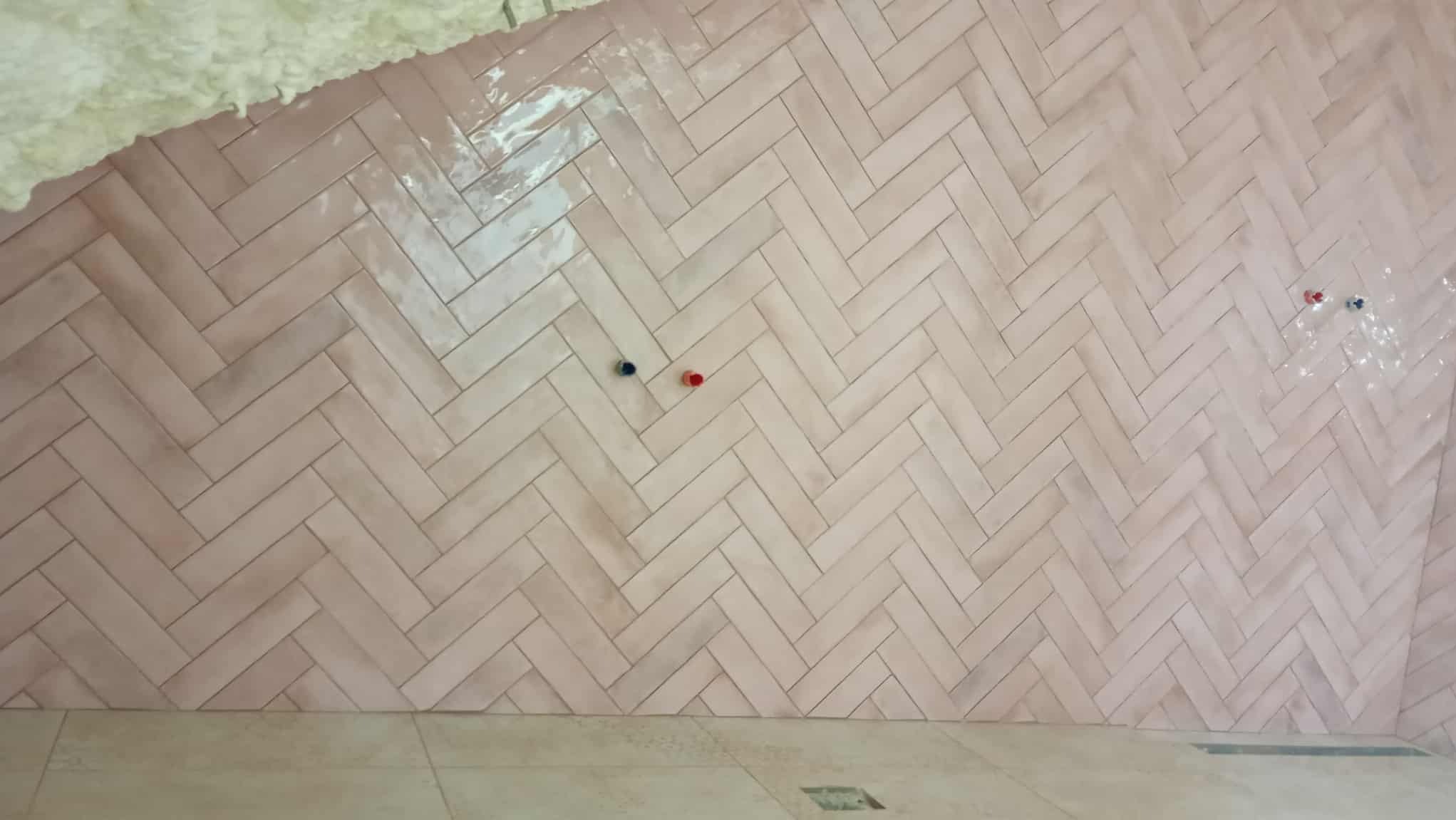 Ściana wyłożona różową, błyszczącą glazurą w jodełkę, z widocznymi przyłączami wodnymi. Fragment sufitu z izolacją i podłoga z płytek w odcieniach beżu.