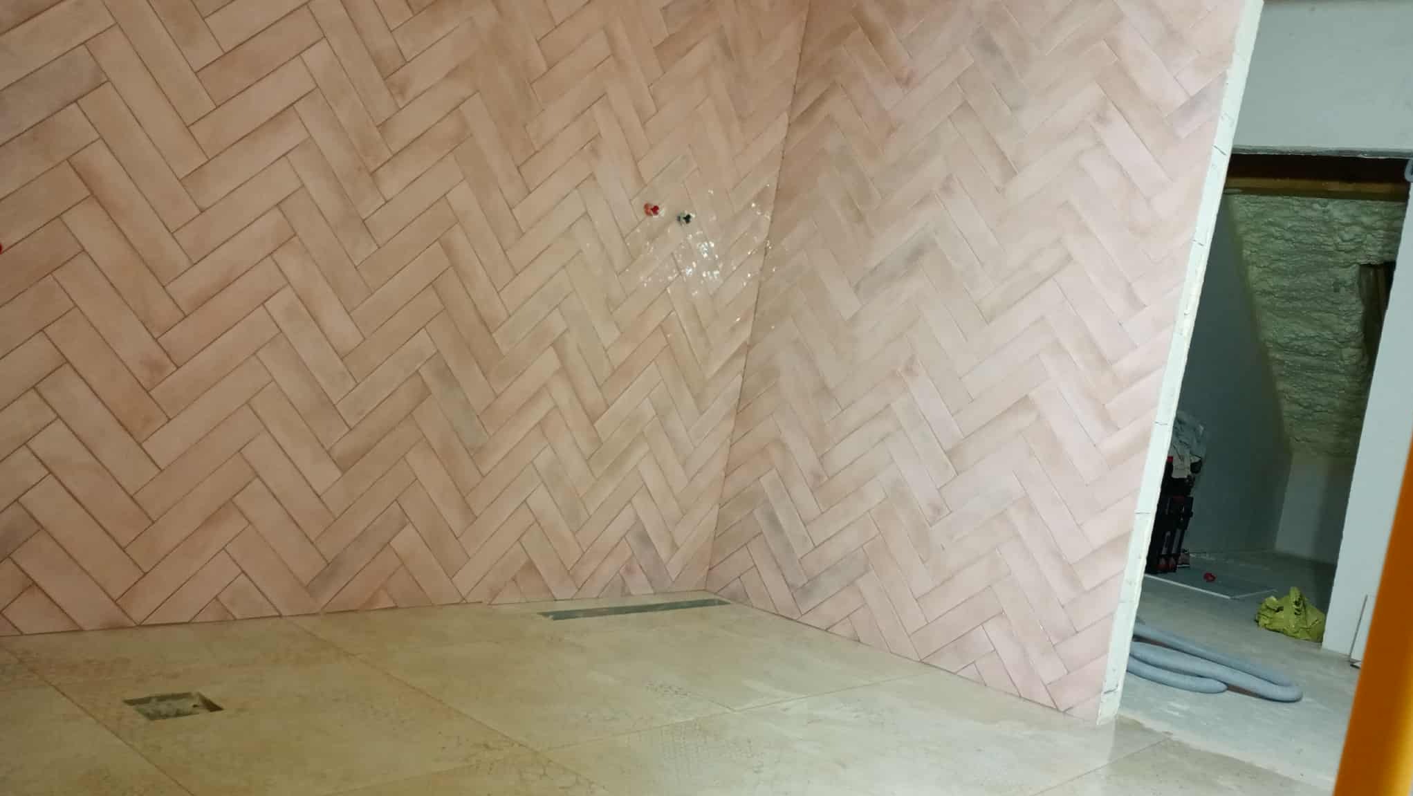 Wykończona łazienka z beżowymi płytkami ułożonymi w jodełkę na ścianach i podłodze. Widoczny odpływ liniowy i instalacja wodna. W tle otwarte drzwi do innego pomieszczenia.