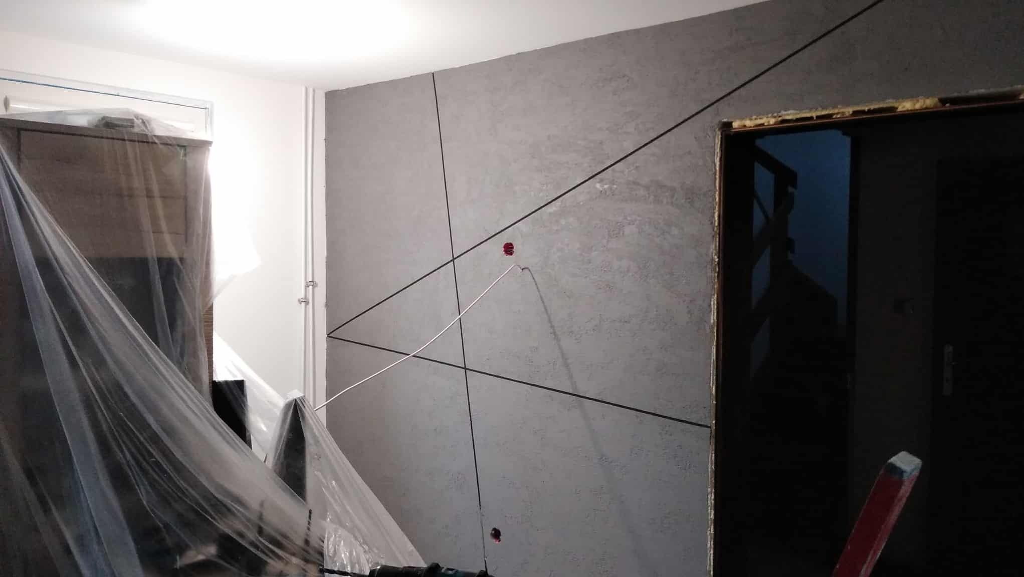 Szara ściana z dekoracyjnymi czarnymi liniami i gniazdkami, częściowo zakryta folią ochronną. Widoczne narzędzia i fragment drzwi.