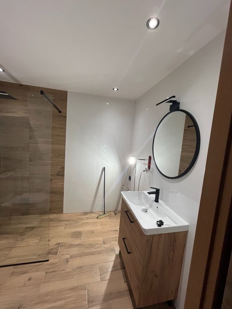 Nowoczesna łazienka z kabiną prysznicową typu walk-in, drewnopodobnymi płytkami i szafką pod umywalkę z czarnym okrągłym lustrem. Minimalistyczny design.