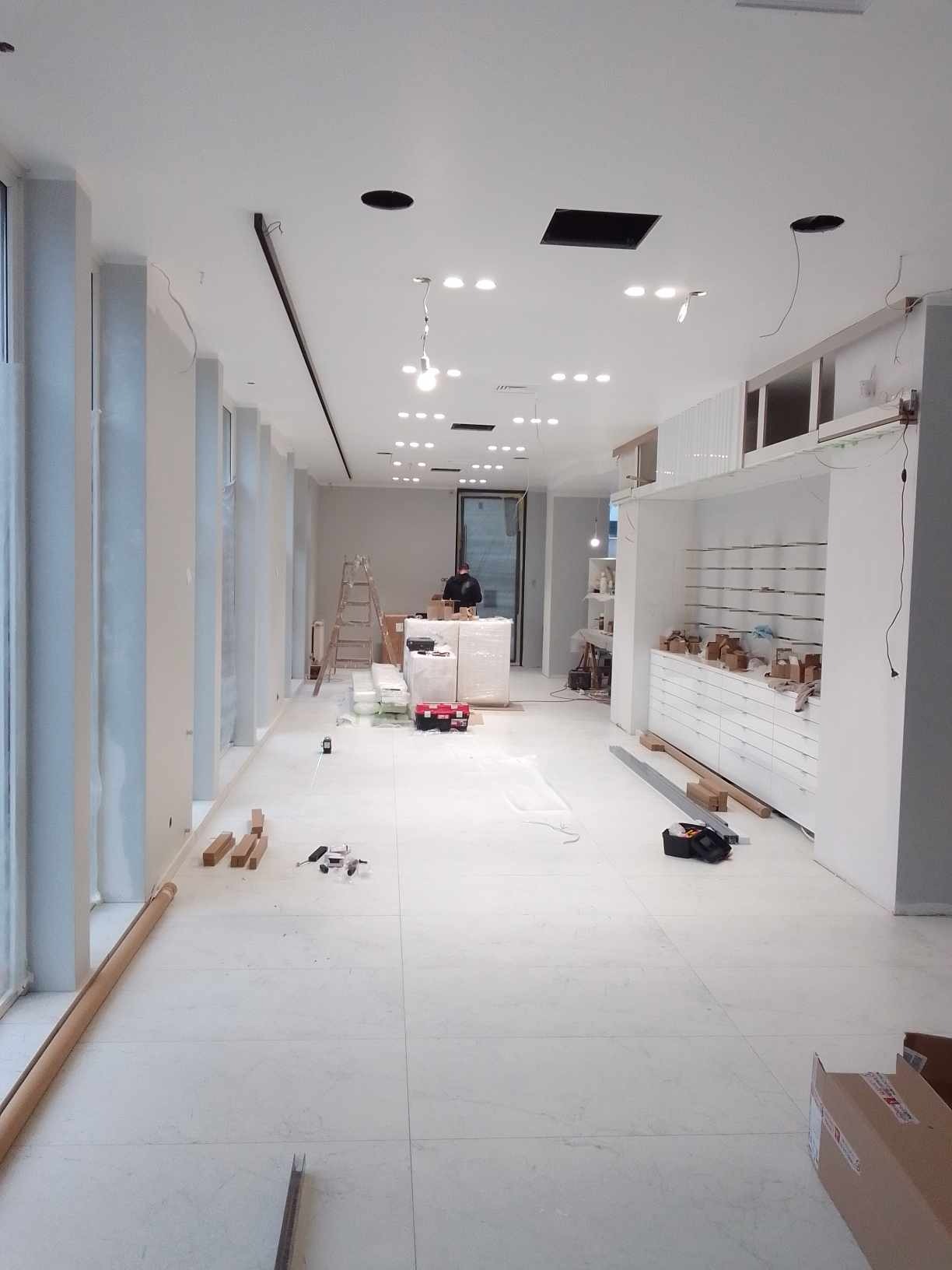 Wnętrze sklepu w trakcie wykańczania: białe ściany, podłoga z płytek, zamontowane regały i oświetlenie. Widoczne narzędzia i materiały budowlane.