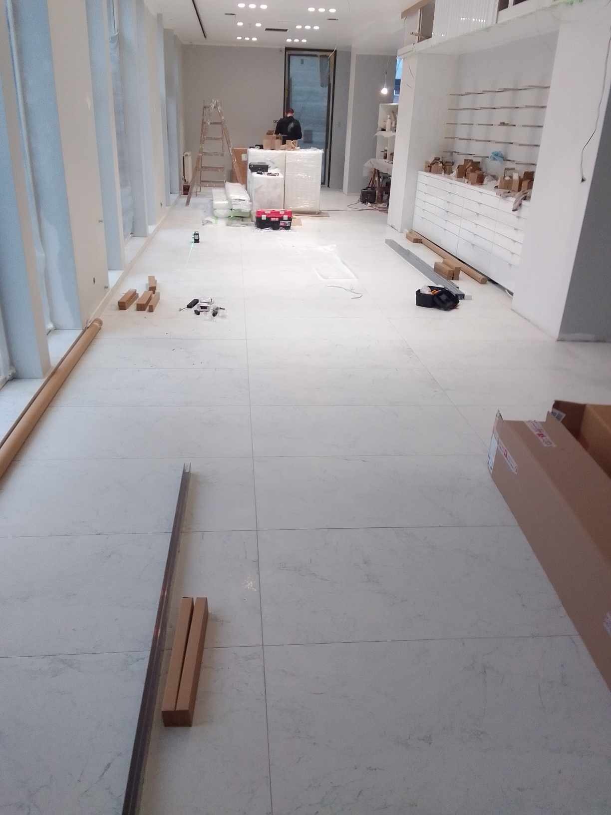 Wnętrze sklepu w trakcie wykańczania: białe płytki podłogowe, regały z szufladami, narzędzia i materiały budowlane. Widoczna drabina i osoba w tle.