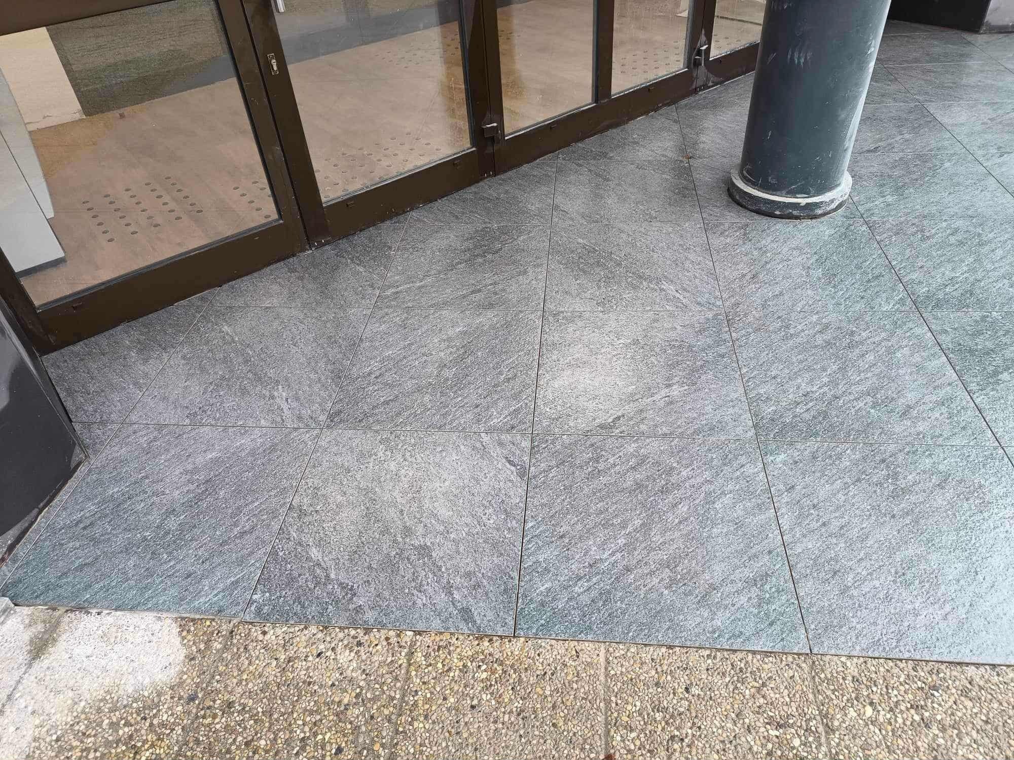 Szare, kwadratowe płytki podłogowe ułożone przy wejściu do budynku w Koszalinie. Widoczny fragment kolumny i drzwi wejściowe z brązową ramą.